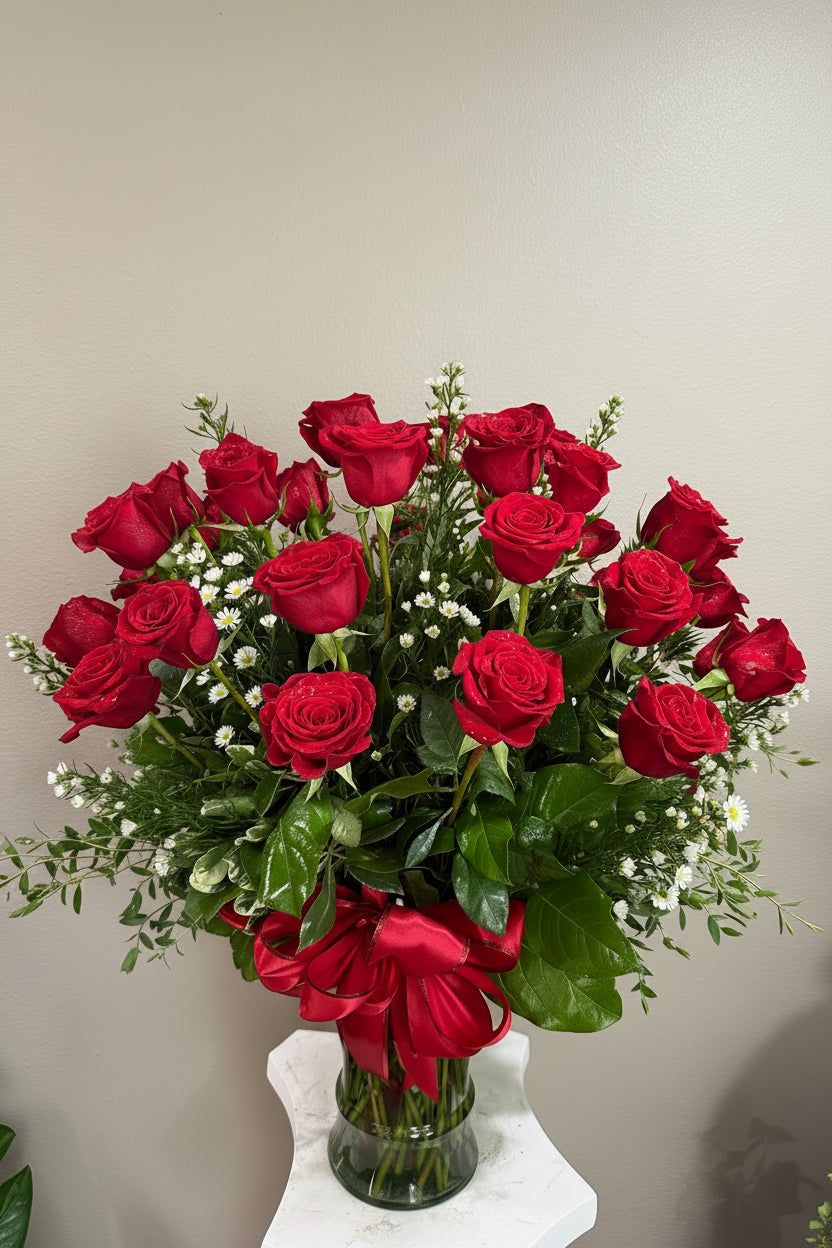 A vibrant bouquet of 24 red roses arranged in a clear glass vase with a large red satin bow, accented with small white daisy-like filler flowers and green foliage,