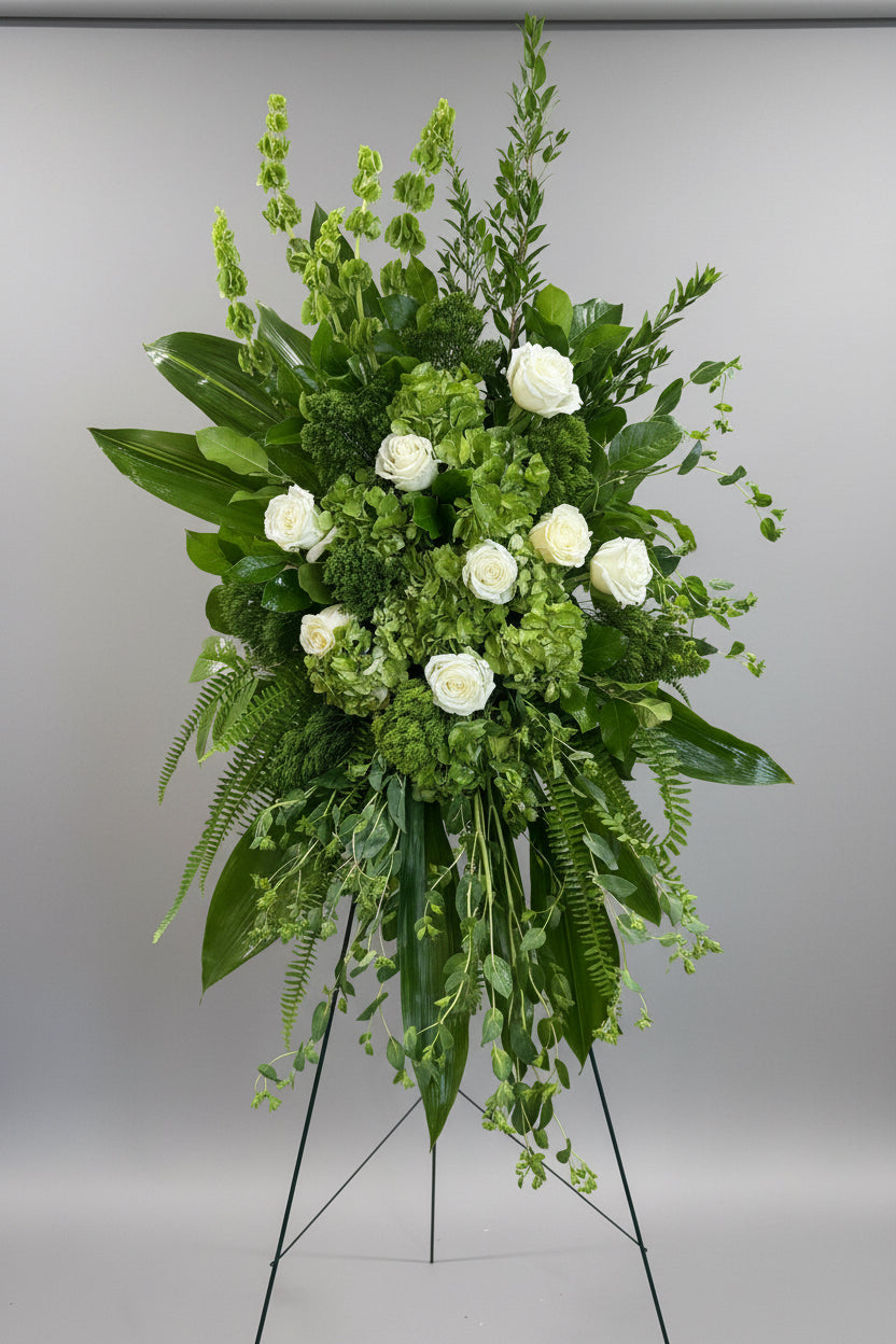 A large green floral arrangement featuring white roses, hydrangeas, ferns, and tall green foliage, displayed on a white pedestal stand in an indoor setting.