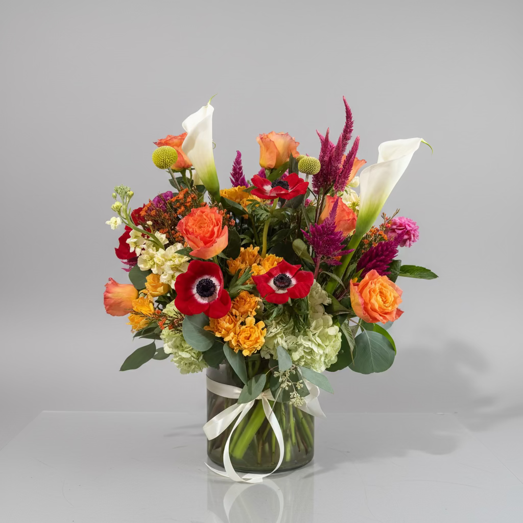 A vibrant mixed floral arrangement in a clear glass vase, featuring red anemones, orange roses, white calla lilies, yellow flowers, and purple ornamental grass, displayed on a wooden stool against a neutral wall.