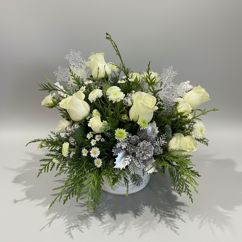 A winter-themed floral arrangement in a white bowl, featuring pale yellow roses, white daisies, green foliage, and silver faux snow elements, set against a gray backdrop.