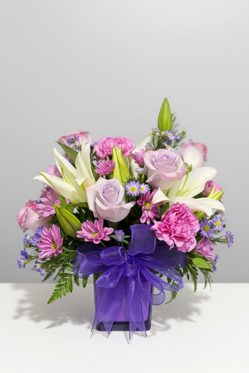 A vibrant floral arrangement featuring white lilies, light purple roses, pink carnations, and purple daisies with green foliage, tied with a large purple ribbon at the bottom.