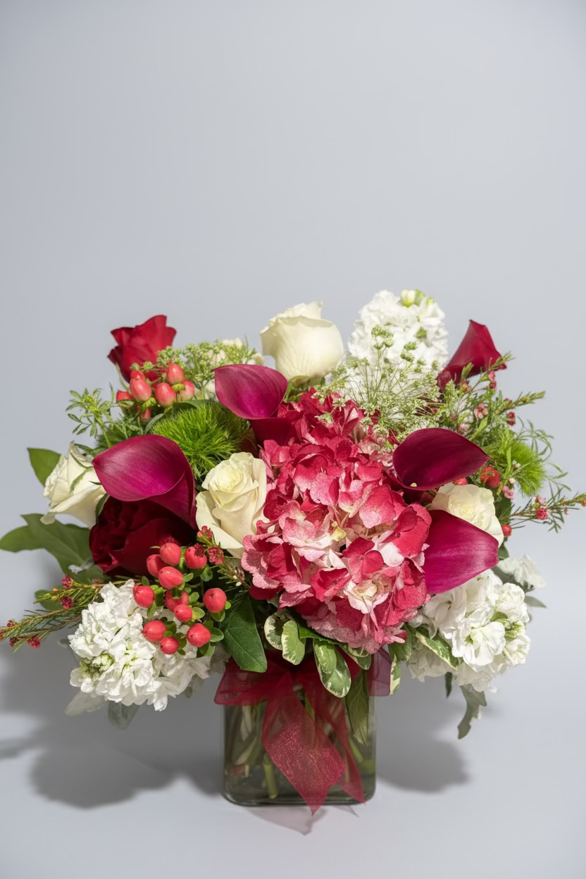 A vibrant floral arrangement in a clear glass vase, featuring bright pink calla lilies, deep pink hydrangeas, white roses, white stock, and green foliage, tied with a pink ribbon against a dark background.