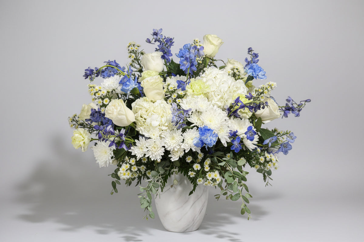 A large floral arrangement featuring white and blue flowers, including roses, chrysanthemums, daisies, and delphiniums, in a textured white vase against a dark background.