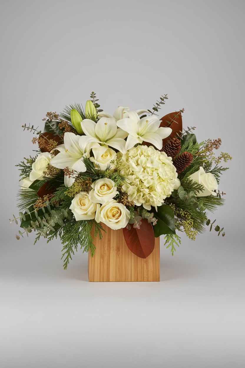 A floral arrangement featuring white lilies, cream roses, and a large cream hydrangea in a light wood box, styled with green foliage, pinecones, and dried botanicals on a dark wooden table.