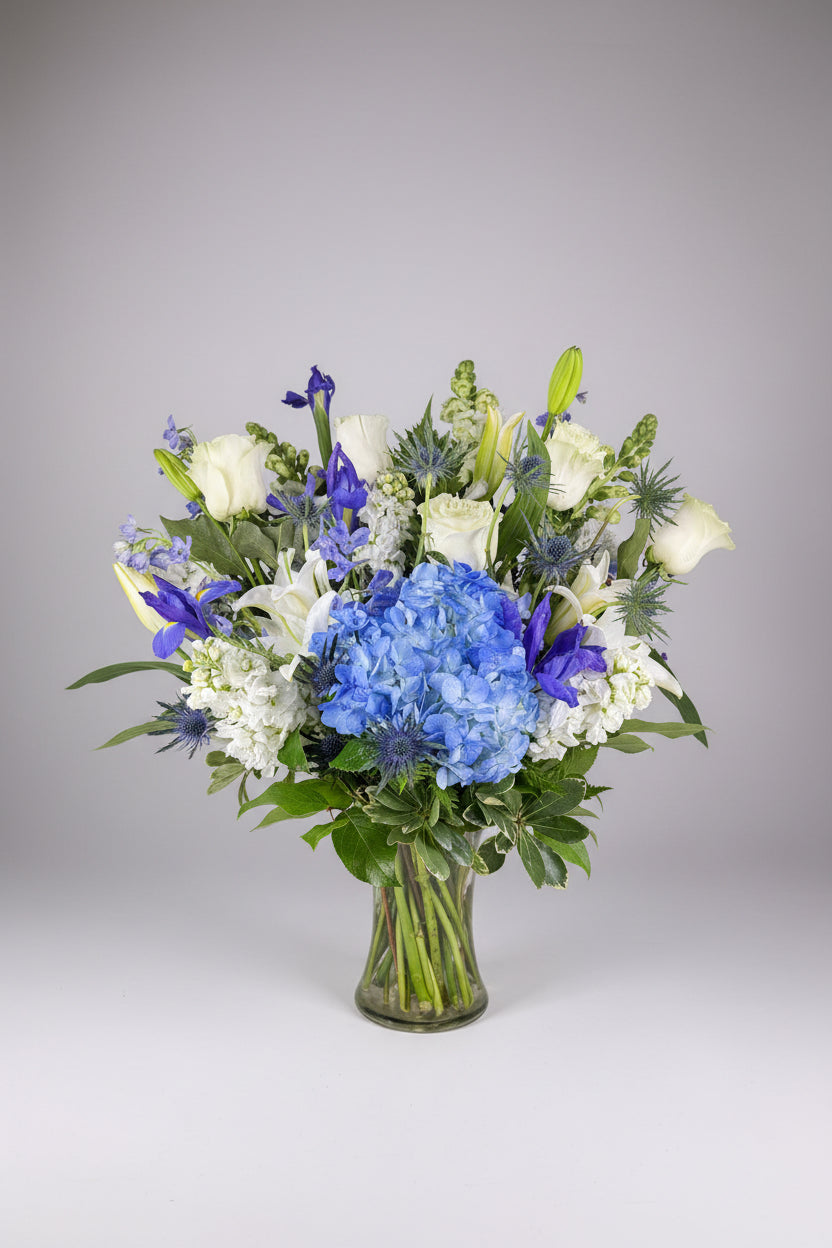 A vibrant flower bouquet in a clear glass vase, featuring a prominent blue hydrangea, white roses, blue irises, and green foliage