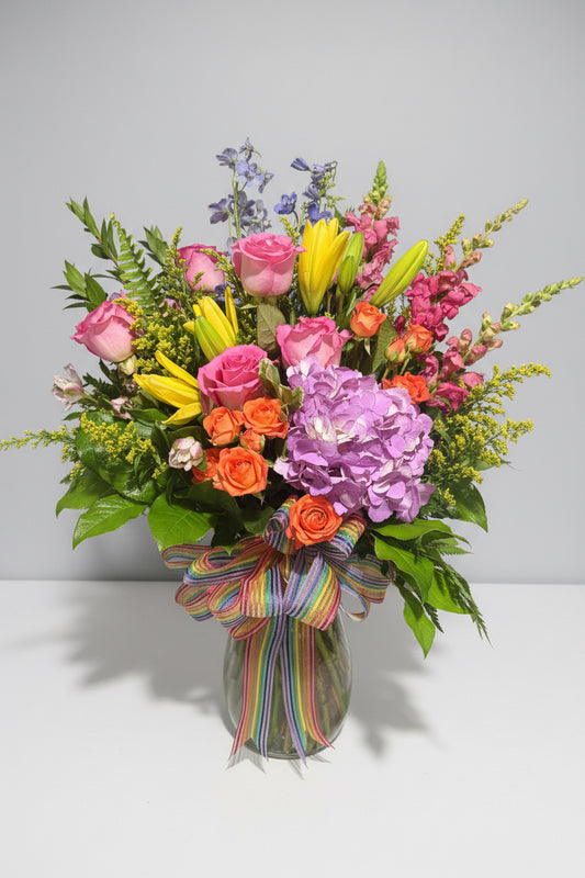 A vibrant floral arrangement featuring pink roses, orange roses, yellow lilies, purple hydrangeas, and green foliage, tied with a rainbow-striped ribbon, shown against a dark background.