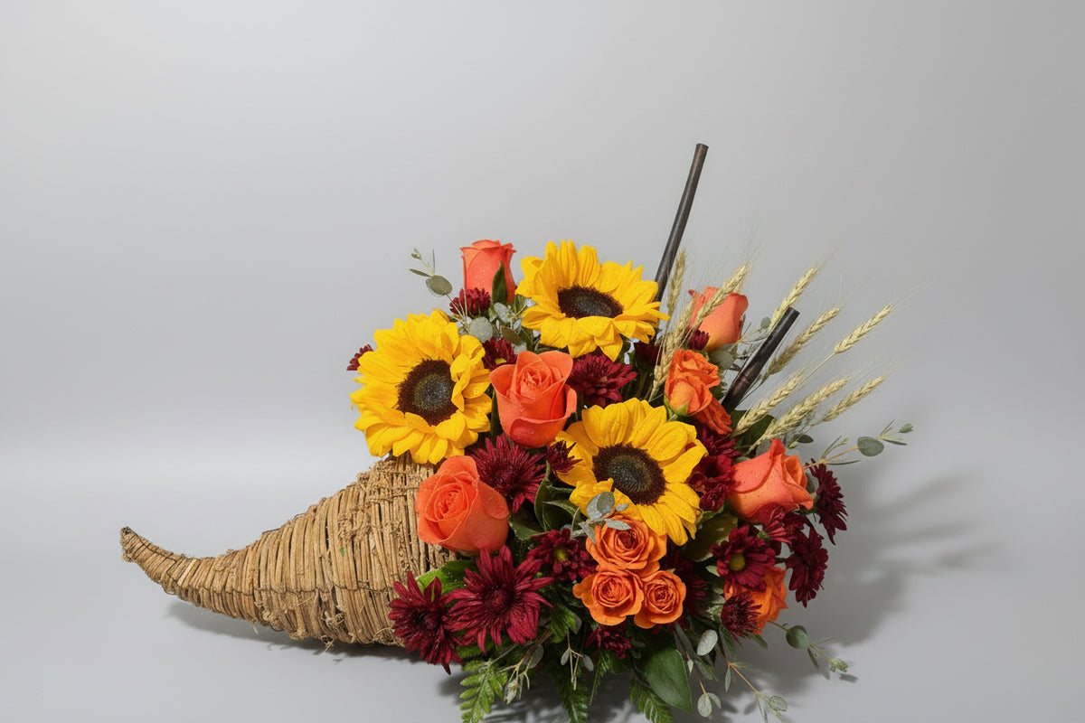 A decorative autumn floral arrangement featuring two large yellow sunflowers, orange and red roses, burgundy chrysanthemums, wheat stalks, and green foliage, styled in a woven cornucopia basket against a dark background.
