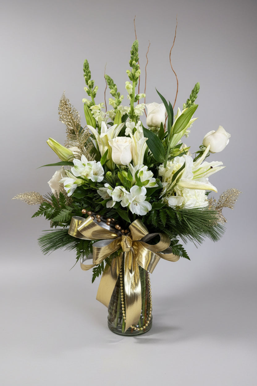 A large floral arrangement in a clear glass vase, featuring white lilies, roses, and white flowers with green foliage, accented by a shiny gold ribbon bow, set against a neutral indoor wall.