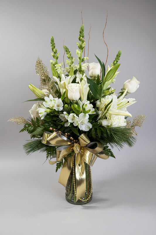 A large floral arrangement in a clear glass vase, featuring white lilies, roses, and white flowers with green foliage, accented by a shiny gold ribbon bow, set against a neutral indoor wall.