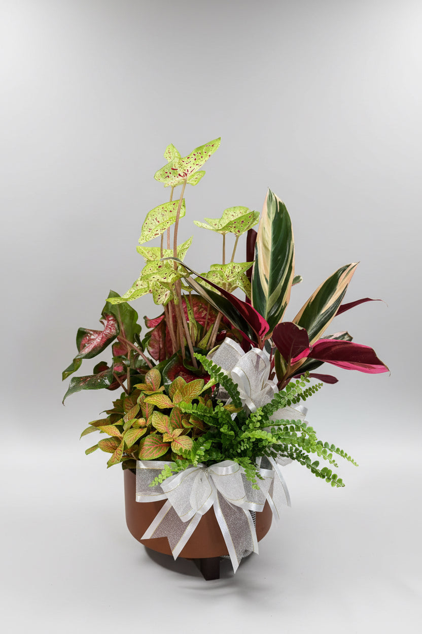 A potted artificial floral arrangement featuring a variety of green, burgundy, and yellow-green foliage plants in a terracotta-colored pot with a white ribbon bow, set against a neutral gray background.