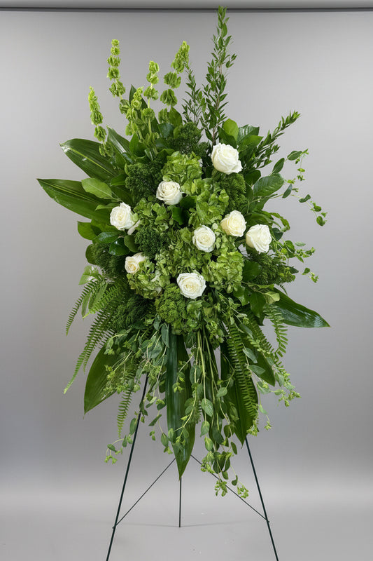 A large green floral arrangement featuring white roses, hydrangeas, ferns, and tall green foliage, displayed on a white pedestal stand in an indoor setting.