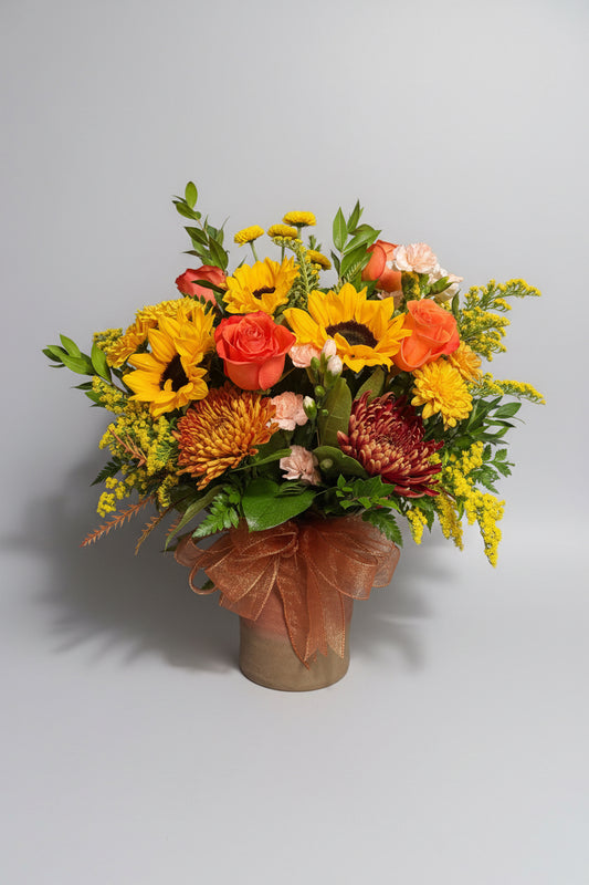 A vibrant autumn floral arrangement featuring yellow sunflowers, orange roses, burgundy chrysanthemums, green foliage, and a brown organza bow, set against a dark black background.