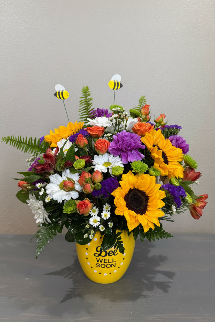 A vibrant artificial flower bouquet featuring large yellow sunflowers, purple and white daisies, orange roses, green ferns, and two bee figurines, arranged in a yellow 'WELL SOON' vase against a gray wall.