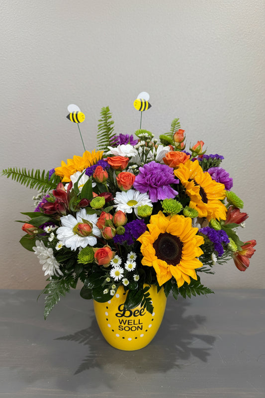 A vibrant artificial flower bouquet featuring large yellow sunflowers, purple and white daisies, orange roses, green ferns, and two bee figurines, arranged in a yellow 'WELL SOON' vase against a gray wall.