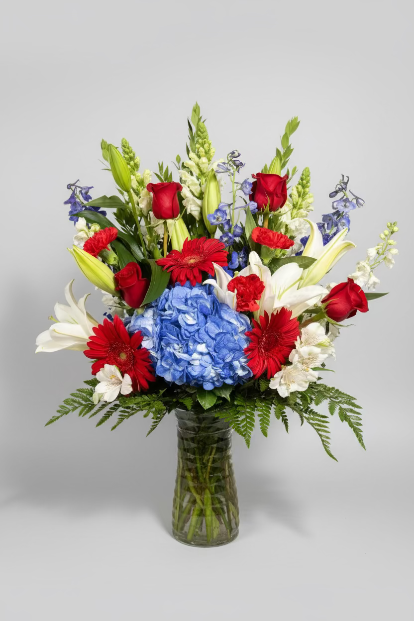 A vibrant mixed flower bouquet in a clear glass vase, featuring a large blue hydrangea, red roses, red gerbera daisies, white lilies, and green foliage against a dark background.