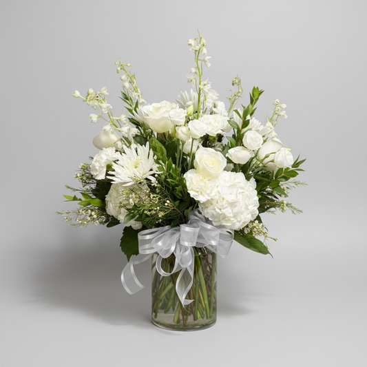 A white floral arrangement featuring roses, hydrangeas, and other white blossoms with green foliage, tied with a sheer white ribbon in a clear glass vase, set against a dark background.