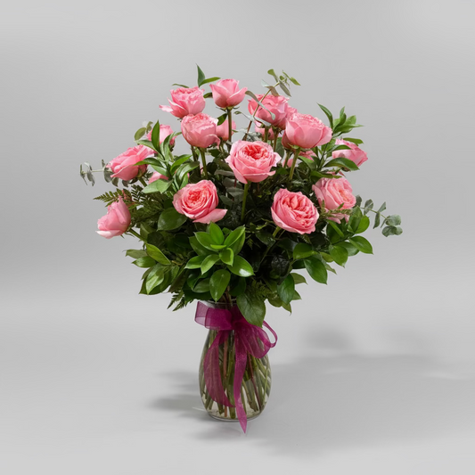 A bouquet of pink roses with green foliage arranged in a clear glass vase tied with a pink ribbon, placed on a white pedestal against a beige wall.