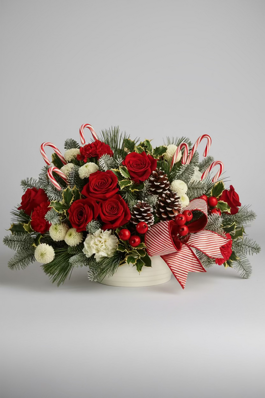 A festive Christmas floral arrangement in a white pot, featuring red roses, white pom-poms, pinecones, candy canes, holly, and a red-and-white striped ribbon, displayed on a reflective table with a softly blurred holiday background.