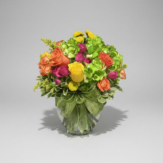 A vibrant mixed flower bouquet in a clear glass vase, featuring orange and yellow roses, magenta blooms, bright yellow button,green hydrangea, solidago and mix greenery