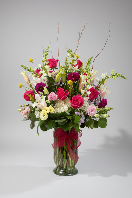 A vibrant floral arrangement in a clear glass vase, featuring pink and red roses, white and yellow lilies, purple flowers, and green foliage