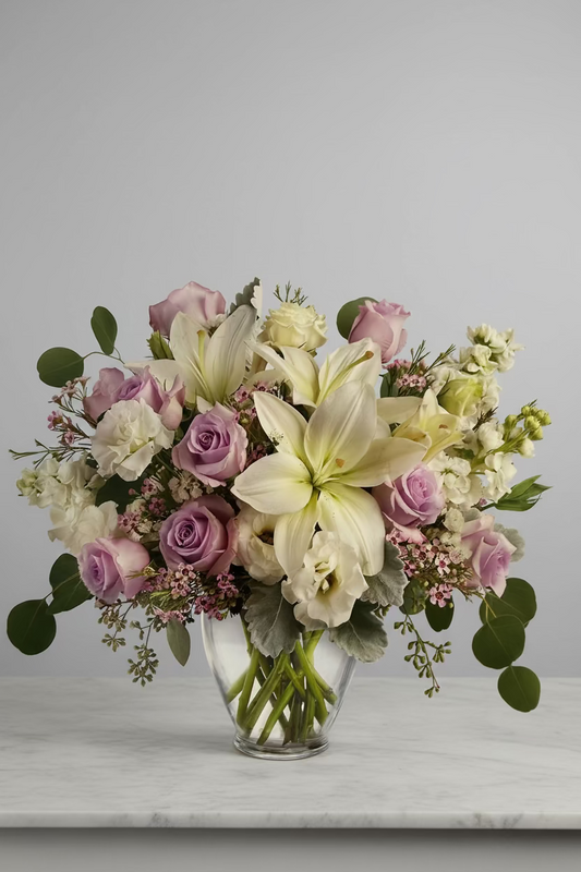 A beautiful floral bouquet in a clear glass vase, featuring large white lilies, light purple roses, white carnations, and green foliage, displayed on a marble tabletop with a soft blue background.