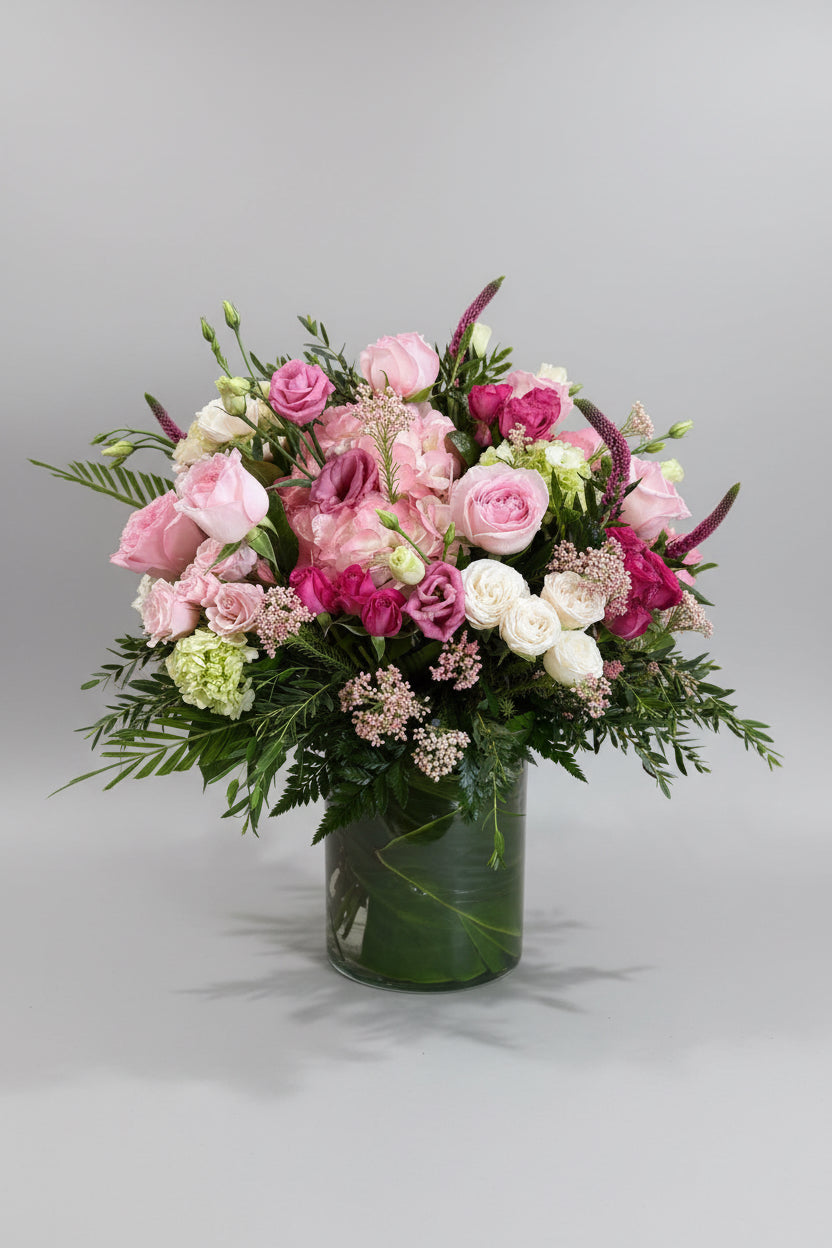 A vibrant floral arrangement featuring pink and white roses, pink lisianthus, and green foliage in a clear glass vase, set on a bright green cloth-covered surface against a neutral beige wall.