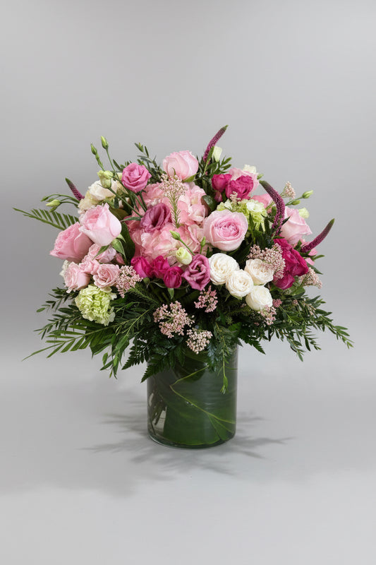 A vibrant floral arrangement featuring pink and white roses, pink lisianthus, and green foliage in a clear glass vase, set on a bright green cloth-covered surface against a neutral beige wall.