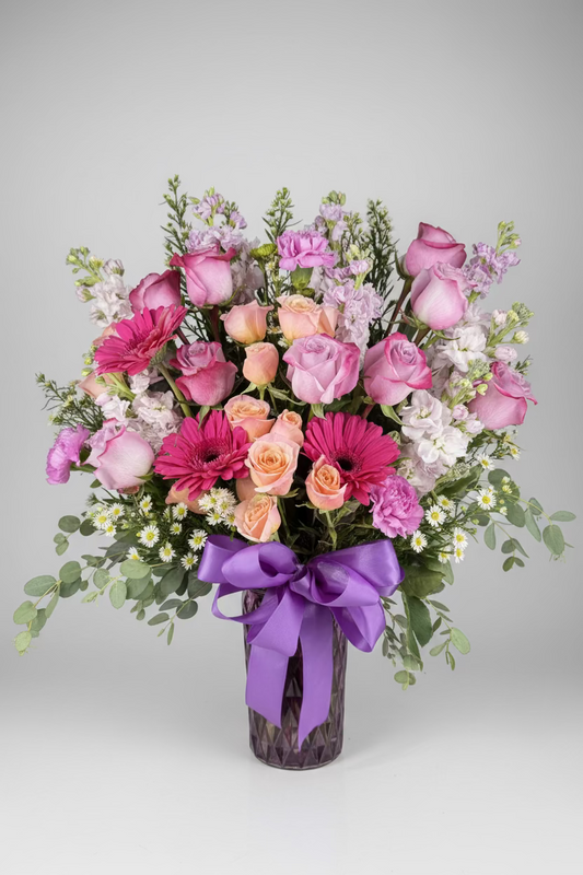 A vibrant floral bouquet featuring pink and orange roses, pink and magenta daisies, purple stock, and green foliage, tied with a large purple ribbon in a glass vase against a dark background.
