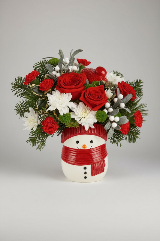 A festive holiday flower bouquet in a snowman-shaped vase, featuring red roses, white chrysanthemums, red carnations, green pine branches, and white berries. The snowman vase is decorated with a red striped hat and a red scarf.