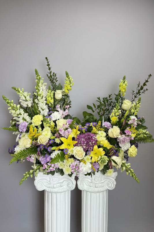 A large floral arrangement featuring yellow lilies, white and yellow roses, purple hydrangeas, and green foliage, displayed in two white classical-style pedestals against a textured gray backdrop.