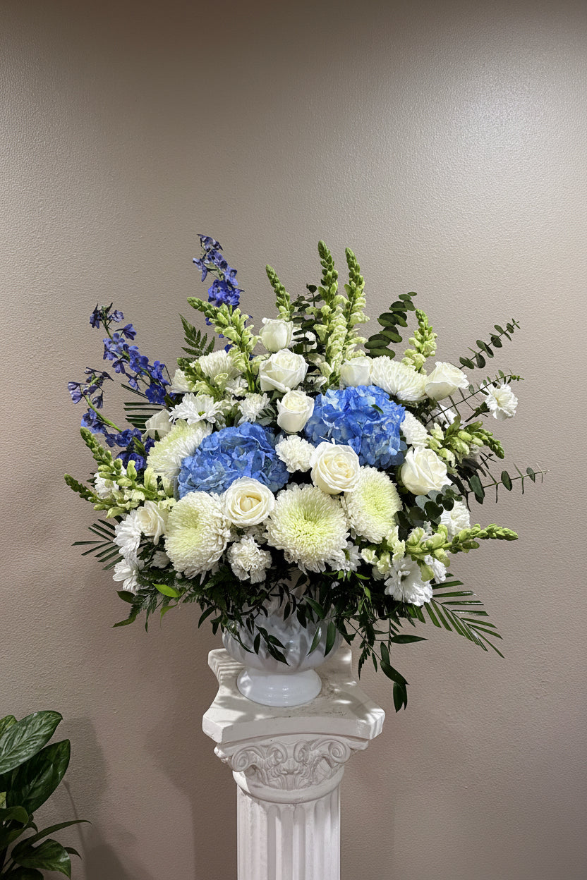 A large floral arrangement in a white pedestal vase, featuring blue hydrangeas, white roses, white chrysanthemums