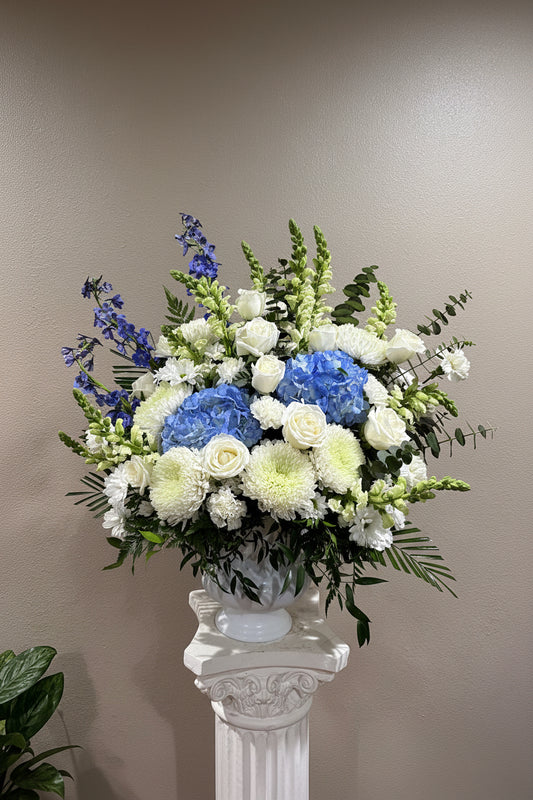 A large floral arrangement in a white pedestal vase, featuring blue hydrangeas, white roses, white chrysanthemums