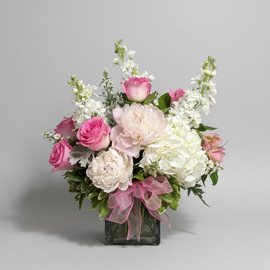 A floral arrangement featuring pink and white peonies, hydrangeas, roses, and white filler flowers in a square glass vase, 
