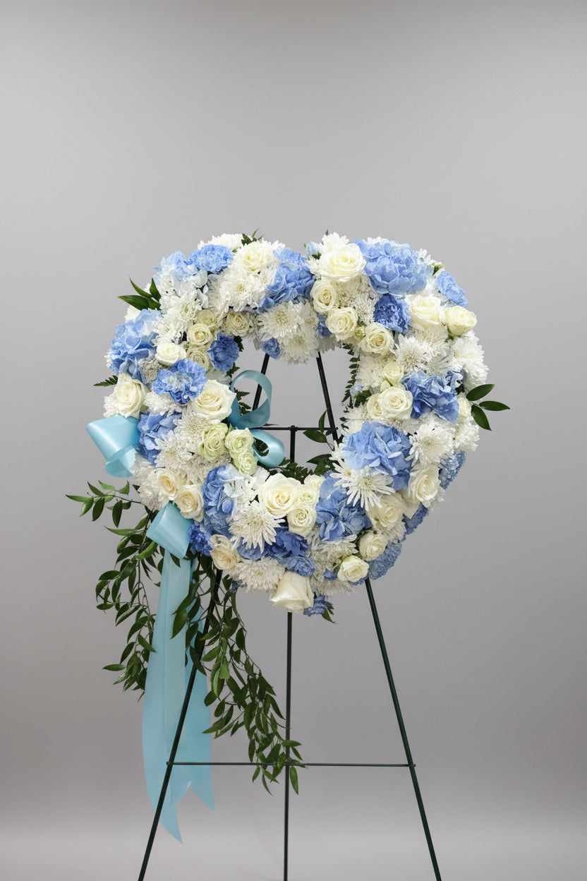 A large heart-shaped floral wreath on a black display stand, decorated with blue and white flowers and a light blue ribbon bow, set against a beige wall.