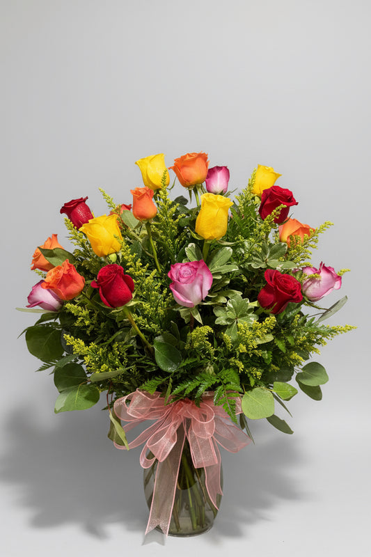 A vibrant mixed flower bouquet featuring red, pink, orange, and yellow roses with green foliage and a sheer pink ribbon, set against a dark background.