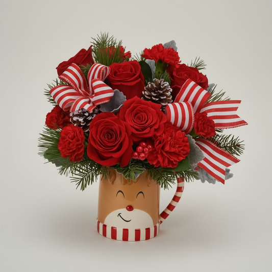 A festive Christmas bouquet in a reindeer mug, featuring red roses, red and white striped ribbons, green pine branches, and small red berries, set against a plain light background.