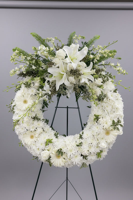 A large floral wreath made of white lilies, daisies, carnations, and green foliage, displayed on a black metal stand in an indoor shop or studio setting.