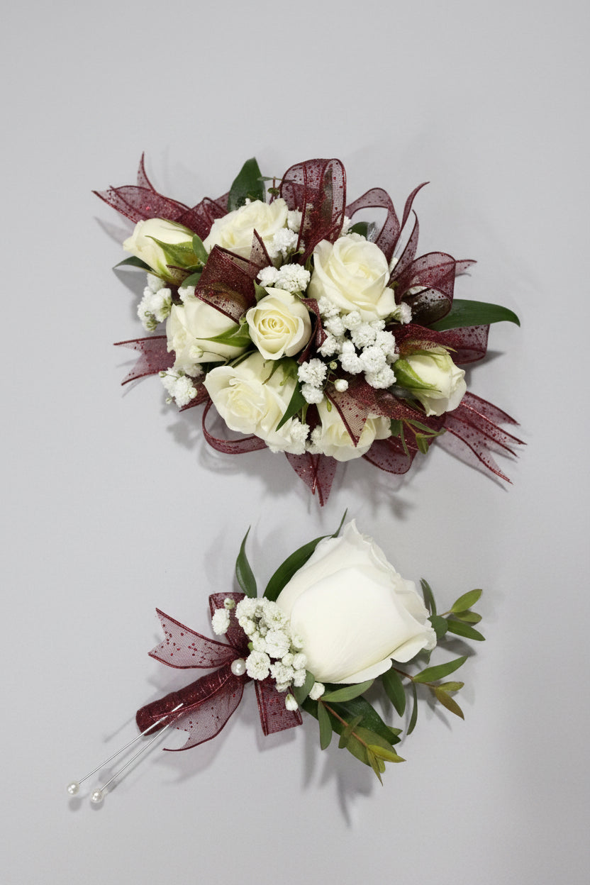 A floral boutonnier and matching bouquet featuring white roses, white baby's breath, green leaves, and dark burgundy sheer ribbon on a light purple fabric background.