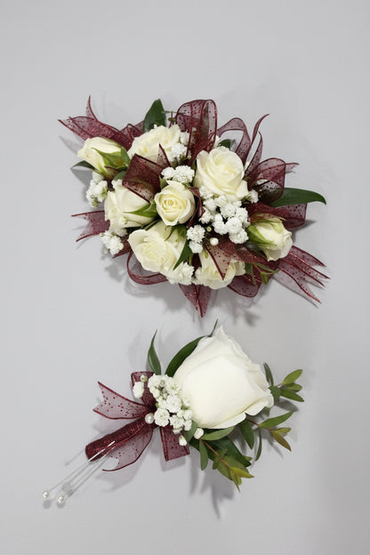 A floral boutonnier and matching bouquet featuring white roses, white baby's breath, green leaves, and dark burgundy sheer ribbon on a light purple fabric background.