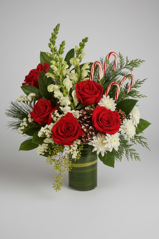 A festive Christmas flower bouquet in a green glass vase, featuring red roses, white chrysanthemums, green sprigs, candy canes, and pinecones, displayed on a wooden tray with small decorated treats against a warm bokeh background.