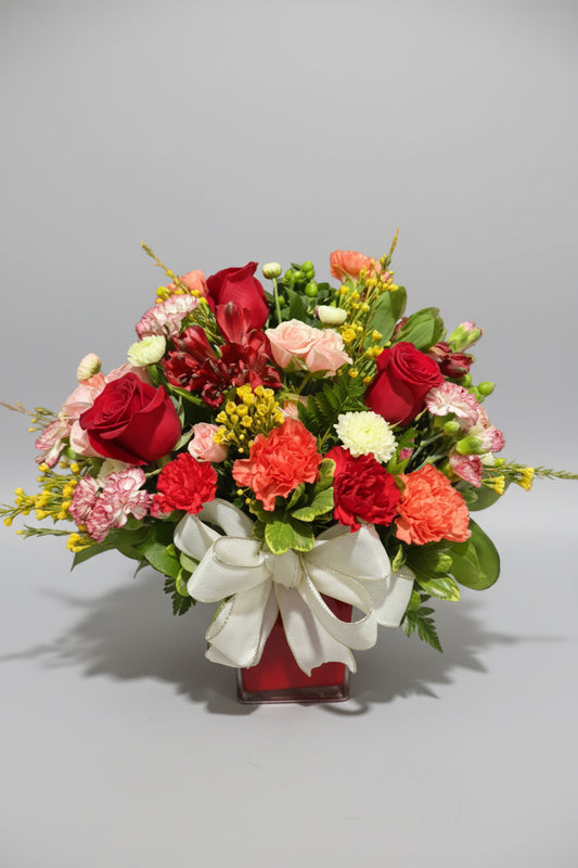 A vibrant floral arrangement featuring red and pink roses, carnations, alstroemeria, and green foliage, tied with a large white ribbon in a square red vase against a dark background.