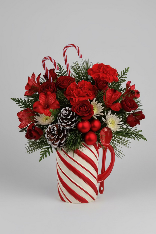 A festive Christmas bouquet in a mug-shaped vase with red and white candy cane stripes, filled with red flowers, green pine branches, white daisies, red ornaments, and candy canes, displayed on a tiled countertop near a window.