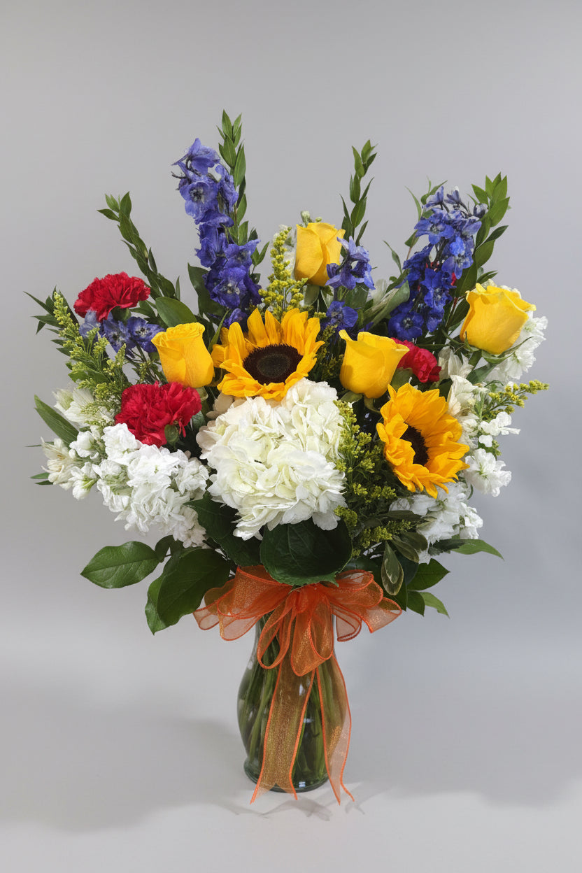 A vibrant mixed flower bouquet in a clear glass vase, featuring yellow sunflowers, yellow roses, blue delphiniums, white hydrangeas, red carnations, and green foliage, tied with an orange ribbon, displayed on a white pedestal against a beige wall.