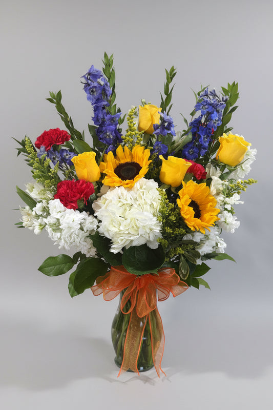 A vibrant mixed flower bouquet in a clear glass vase, featuring yellow sunflowers, yellow roses, blue delphiniums, white hydrangeas, red carnations, and green foliage, tied with an orange ribbon, displayed on a white pedestal against a beige wall.