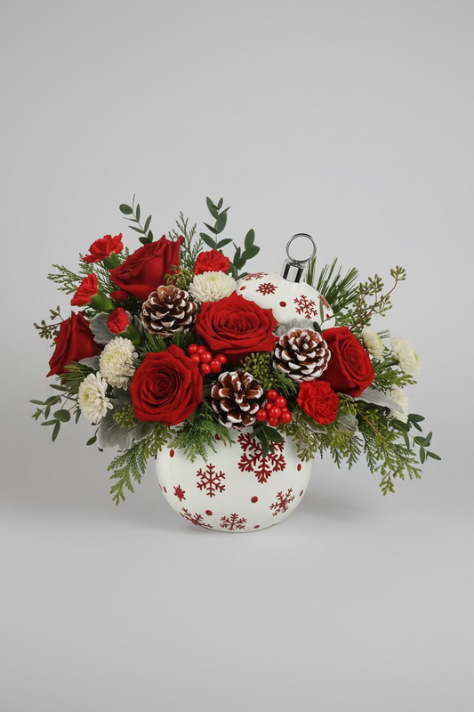 A festive holiday bouquet in a white ceramic pot decorated with red snowflakes, filled with red roses, white chrysanthemums, pinecones, and green foliage, set against a white background.